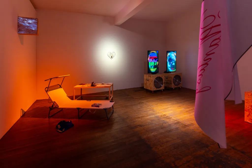 An hot orange lit room, with water projection on chip board, deckchair to lounge on, books to the side. A white heart sculpture is affixed on the back wall, to the right 2 screens with spiralling graphics. To the front right,  a white flag hangs 