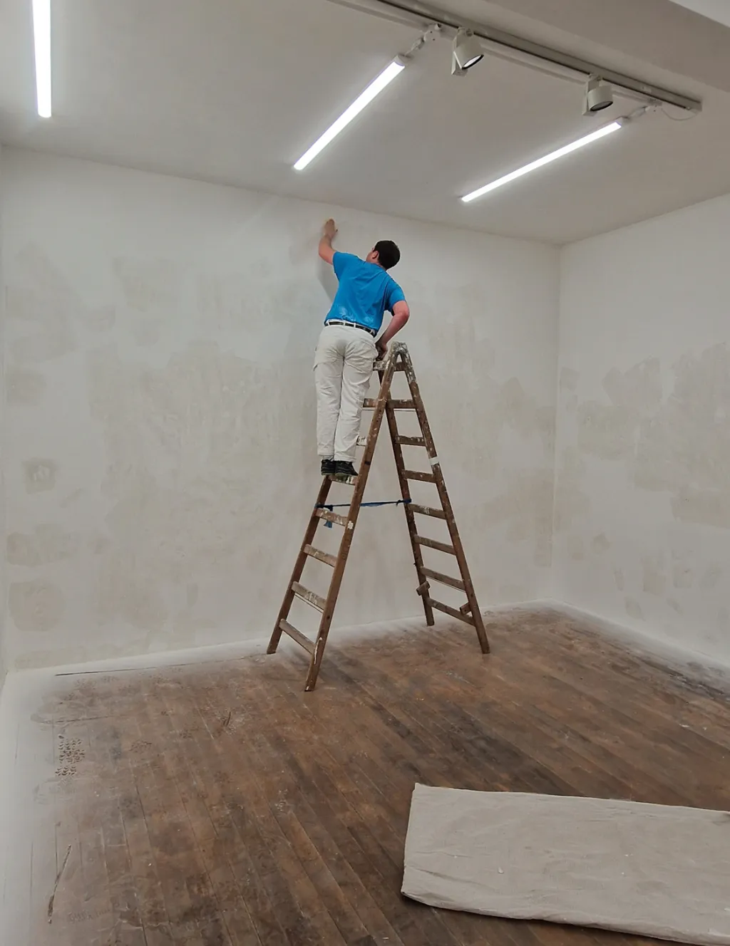 Image of a man standing on a ladder, sanding a gallery wall in preparation for painting it.